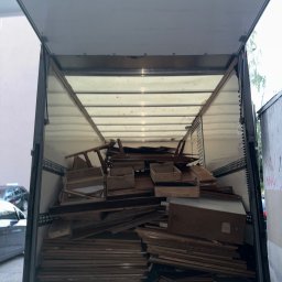 ProTrans - Wnętrze otwartego busa wypełnione elementami mebli z ciemnego drewna, prawdopodobnie po demontażu lub przygotowane do transportu. Widok z tyłu pojazdu.