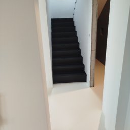 Fajne Podłogi - Jasna, jednolita podłoga i czarne schody w minimalistycznym wnętrzu. Widoczny surowy otwór drzwiowy i fragment ściany z otworem na gniazdko.