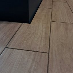 Dawid Panek - Ułożone płytki podłogowe imitujące drewno, przylegające do ciemnej ściany. Widoczna fuga i struktura płytek. Ujęcie z niskiego kąta.