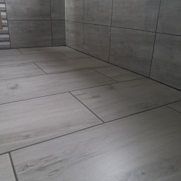 Dawid Panek - Wykończona łazienka z szarymi, drewnopodobnymi płytkami na podłodze i ścianach. Widoczny narożnik pomieszczenia i fugi. Minimalistyczny design.