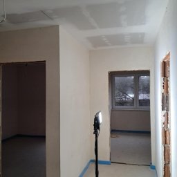 Dawid Panek - Wnętrze w trakcie prac wykończeniowych: ściany po szpachlowaniu, sufit z widocznymi łączeniami płyt, zabezpieczone podłogi, oświetlenie budowlane.