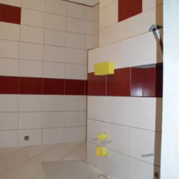 Wnętrze łazienki w trakcie remontu z częściowo położonymi białymi i bordowymi płytkami, widoczne żółte rury i narzędzia glazurnicze na podłodze.