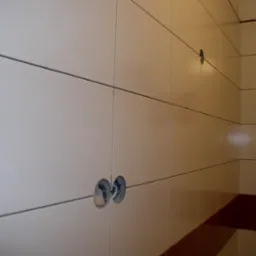 Ściana wyłożona białymi, prostokątnymi płytkami z ciemnymi fugami, z wywierconymi otworami na instalacje, uzupełniona bordiurą w kolorze bordowym.