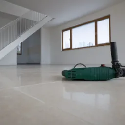 Wnętrze w trakcie remontu: widoczne płytki podłogowe w odcieniach beżu, drabina schodowa, okno z drewnianą ramą, wiertarka udarowa i zwinięty przewód elektryczny na posadzce.