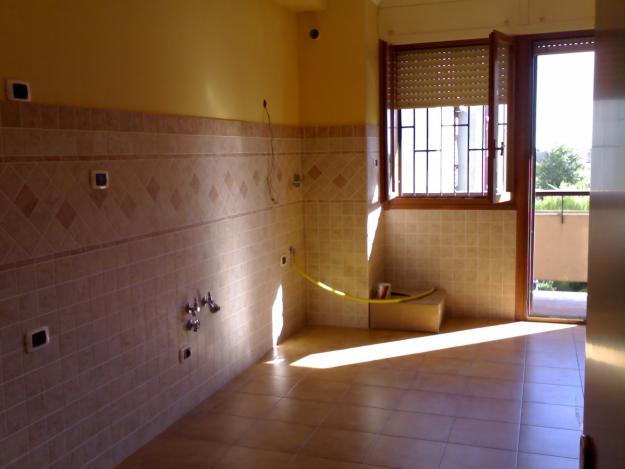 Wnętrze kuchni w trakcie remontu: beżowe płytki ścienne z dekoracyjnym pasem, przygotowane przyłącza wodne, żółty przewód leżący na podłodze z płytek, okno z roletą i widok na balkon.