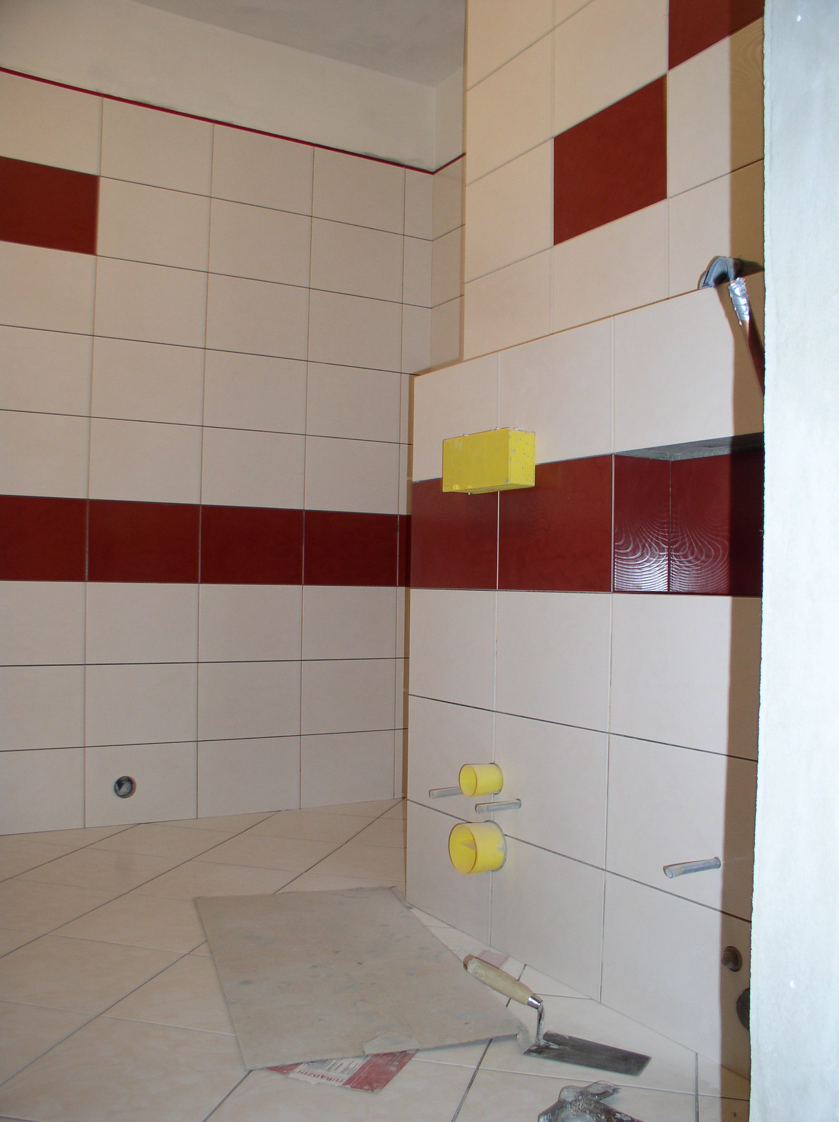Wnętrze łazienki w trakcie remontu z częściowo położonymi białymi i bordowymi płytkami, widoczne żółte rury i narzędzia glazurnicze na podłodze.
