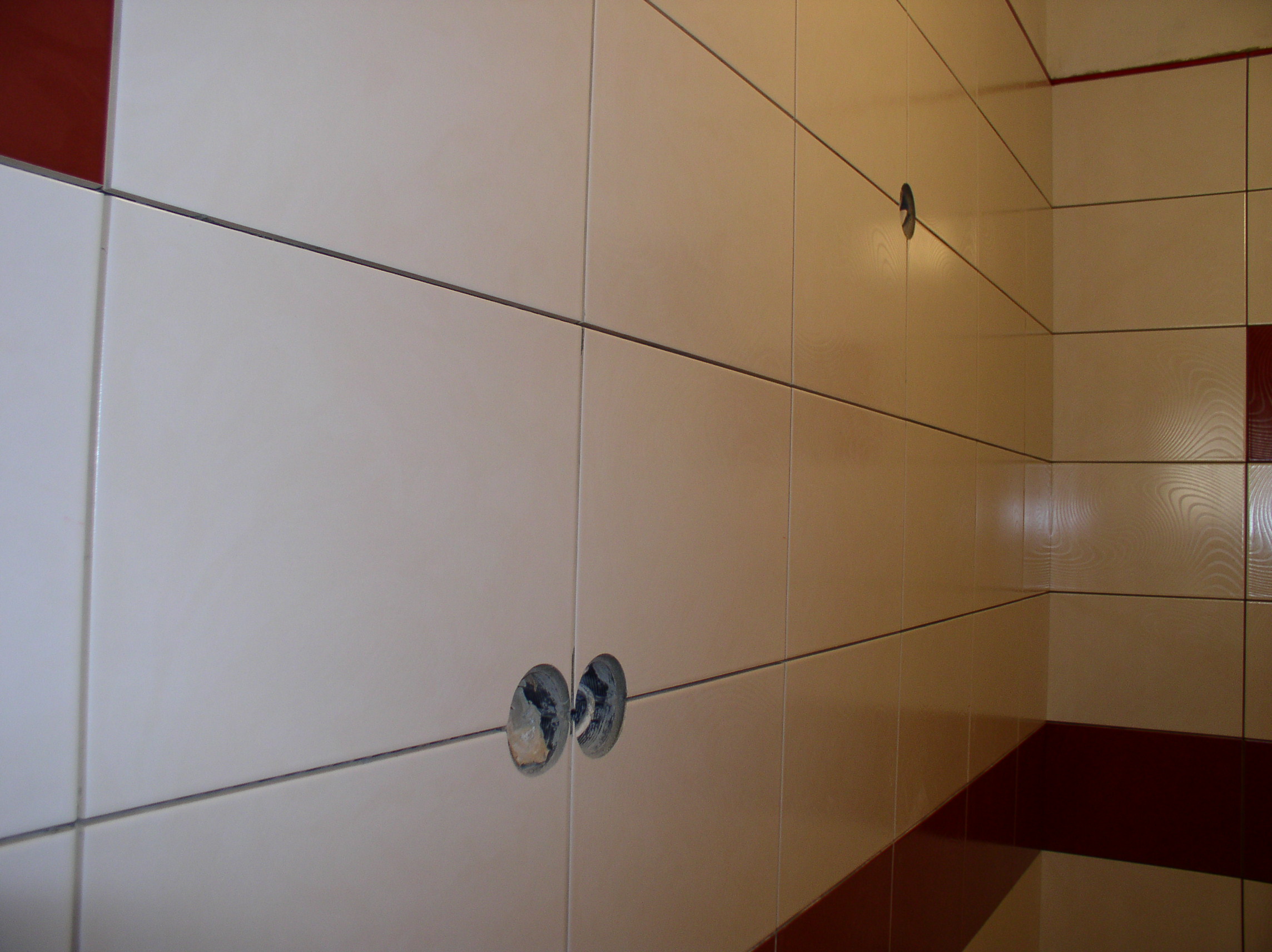 Ściana wyłożona białymi, prostokątnymi płytkami z ciemnymi fugami, z wywierconymi otworami na instalacje, uzupełniona bordiurą w kolorze bordowym.