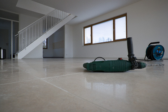 Wnętrze w trakcie remontu: widoczne płytki podłogowe w odcieniach beżu, drabina schodowa, okno z drewnianą ramą, wiertarka udarowa i zwinięty przewód elektryczny na posadzce.