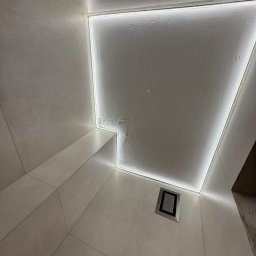 Wojciech Maćkowiak firma handlowo-usługowa - Nowoczesna łazienka z podświetlanym sufitem LED, płytki w odcieniach beżu, minimalistyczny design, kratka wentylacyjna w rogu. Nietypowy kąt ujęcia.