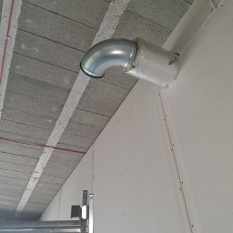 Thermiq - Montaż rury wentylacyjnej, izolowanej folią, wystającej ze ściany w surowym wnętrzu. Widoczny fragment sufitu z betonowych płyt i drabina monterska.