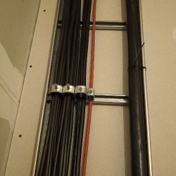 Marcel Konicki - Ułożone kable elektryczne w korycie na ścianie, zabezpieczone uchwytami. Widoczne czarne i czerwone przewody. Koryto przymocowane do ściany z płyt gipsowych.