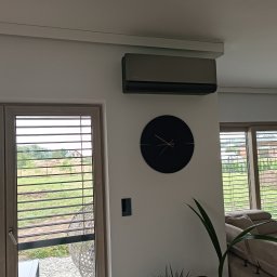RAF-MART - Szary klimatyzator ścienny w nowoczesnym salonie z zegarem i widokiem na zieleń przez okno z żaluzjami. Minimalistyczny design, stonowana kolorystyka.