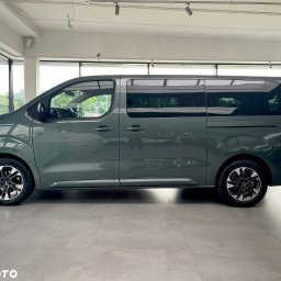 VP Rent Bus - Szary, przeszklony van z ciemnymi felgami, zaparkowany w jasnym salonie. Widok z boku, zarys drugiego pojazdu w tle. Długi rozstaw osi.
