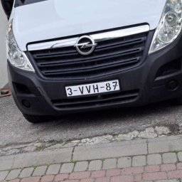 Wojciech chachura - Biały bus marki Opel Master z widoczną tablicą rejestracyjną, zaparkowany na brukowanej nawierzchni. Ujęcie z dołu, podkreślające wysokość pojazdu.