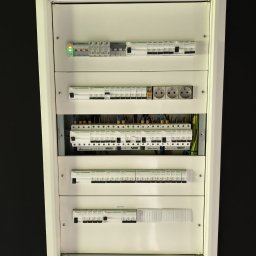 Usługi Elektryczne Maciej Krasnodębski - Rozdzielnica elektryczna w białej obudowie, widoczne zabezpieczenia, gniazda i przewody. Ujęcie z bliska, na czarnym tle ściany.