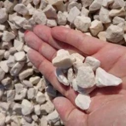 Dłoń trzymająca garść białych kamieni ozdobnych na tle sterty takich samych kamieni.