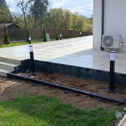 Firma handlowo usługowa Piotr Kozieł - Nowoczesny taras z jasnych płyt, oświetlony czarnymi lampami. Widoczne schody i klimatyzator na ścianie budynku. Tło: zielony trawnik i drzewa.