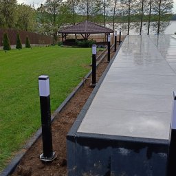 Firma handlowo usługowa Piotr Kozieł - Instalacja oświetlenia zewnętrznego: czarne słupki z białymi kloszami wzdłuż ścieżki z szarych płyt, w tle altana i jezioro. Widok na ogród.
