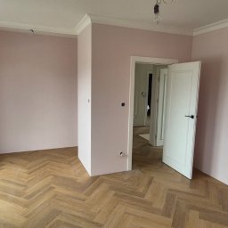 Michał Kowalski - Jasny pokój po remoncie: parkiet w jodełkę, świeżo pomalowane ściany na różowo, białe listwy przysufitowe i drzwi. Widoczne gniazdka elektryczne.