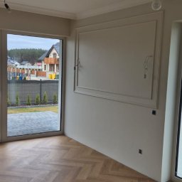 Michał Kowalski - Jasne wnętrze z parkietem w jodełkę, ozdobną sztukaterią na ścianie i dużymi oknami z widokiem na ogród. Widoczne niedokończone instalacje elektryczne.