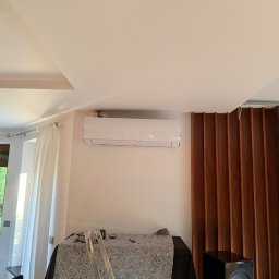 MBTklima - Biały klimatyzator zamontowany na ścianie w nowoczesnym wnętrzu. Widoczne narzędzia i drabina sugerują niedawny montaż. Okno z widokiem na zieleń w tle.