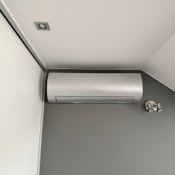 MBTklima - Zamontowany srebrny klimatyzator ścienny marki Sinclair na szarej ścianie w pomieszczeniu ze skośnym sufitem i oświetleniem punktowym.