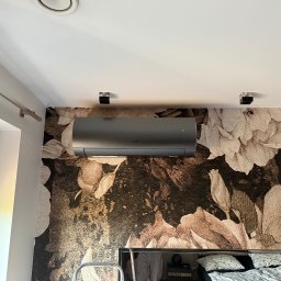 MBTklima - Szary klimatyzator zamontowany na ścianie z tapetą w kwiaty nad łóżkiem. Widoczne lampy sufitowe i fragment okna. Minimalistyczny design.