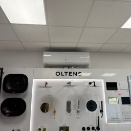 MBTklima - Biała klimatyzacja ścienna nad ekspozycją armatury łazienkowej marki Oltens w sklepie. Widoczne umywalki, baterie i panele WC w różnych kolorach.