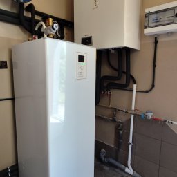 SW-INSTAL - Nowoczesna instalacja grzewcza z białą pompą ciepła stojącą i wiszącym kotłem gazowym Termet, z niebieskim naczyniem wzbiorczym i rurami w pomieszczeniu technicznym.