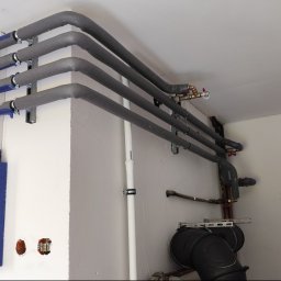 SW-INSTAL - Szereg zaizolowanych rur grzewczych lub wodnych, mocowanych do białej ściany i sufitu, z widocznymi zaworami i skrzynką elektryczną, w pomieszczeniu technicznym.