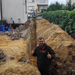 SW-INSTAL - Uśmiechnięty pracownik w czarnej bluzie, stojący w głębokim wykopie ziemnym, pokazuje kciuki w górę. W tle mini-koparka i stosy kostki brukowej obok budynku.