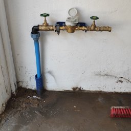 SW-INSTAL - Nowo zamontowana mosiężna instalacja wodomierza z zielonymi zaworami i niebieską rurą, osadzona na białej ścianie nad mokrą, zabrudzoną posadzką.