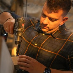 ElektrykLubartow24 - Mężczyzna w kraciastej koszuli wierci w drewnianym elemencie wiertarką akumulatorową. Widoczny zegarek na ręce i oświetlenie warsztatowe.