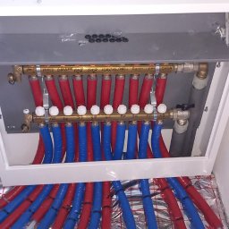 UBS Usługi Budowlane Stępień - Rozdzielacz ogrzewania podłogowego w białej szafce, z widocznymi czerwonymi i niebieskimi rurami. Instalacja na folii izolacyjnej, czarna rura w tle.