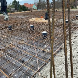 ELTOM Tomasz Leśniewski - Zbrojenie fundamentów domu jednorodzinnego z widocznymi pionowymi prętami i elementami instalacji kanalizacyjnej. W tle zabudowa mieszkalna i pochmurne niebo.