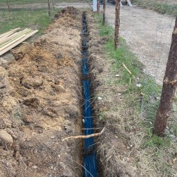PRIME SERVICE - Wkopane w ziemię niebieskie rury, prawdopodobnie peszle, w wykopie wzdłuż ogrodzenia z siatki. W tle widoczny sprzęt budowlany i teren zielony.