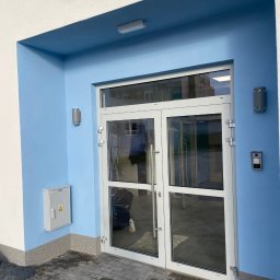 PRIME SERVICE - Błękitne wejście do budynku z białą stolarką okienną i drzwiową. Na ścianie skrzynka elektryczna i lampy zewnętrzne. Widoczny domofon i kostka brukowa.