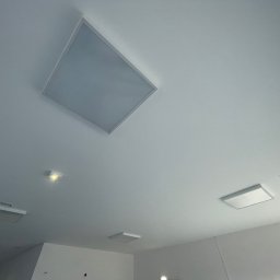 PRIME SERVICE - Biały sufit z zamontowanymi panelami LED o kwadratowym kształcie i okrągłymi otworami wentylacyjnymi. Ujęcie z dołu, widoczne elementy instalacji.