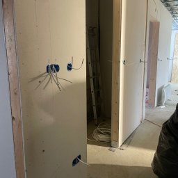 PRIME SERVICE - Instalacja elektryczna w nowym budynku: widoczne kable wystające ze ścian, puszki instalacyjne, drabina w tle. Etap prac przed wykończeniem.