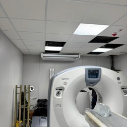 Comfort Flow - Biała klimatyzacja kasetonowa zamontowana w suficie podwieszanym pomieszczenia z tomografem komputerowym Optima. Widoczne elementy instalacji i oświetlenia.