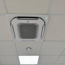 Comfort Flow - Kasetonowa klimatyzacja sufitowa z zielonym wyświetlaczem, zamontowana w biurze z rastrowym sufitem podwieszanym i okrągłymi nawiewnikami.
