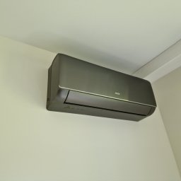 Comfort Flow - Szary klimatyzator AUX zamontowany na jasnej ścianie w rogu pomieszczenia, widoczny fragment sufitu. Minimalistyczny design, nowoczesne wykończenie.