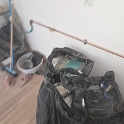 Transporcik - Wnętrze po remoncie: worki na śmieci, wiadro z narzędziami, mop i zmiotka przy ścianie z instalacją. Widoczne ślady prac wykończeniowych.