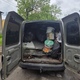 Transporcik - Otwarty bagażnik busa wypełniony workami, wiadrami po farbach i innymi przedmiotami. Widoczne otwarte drzwi i otoczenie budynku z zielenią.