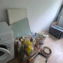 Transporcik - Prowizoryczny składzik materiałów budowlanych: worki z zaprawą, płytki, wiaderka z farbą, grzejnik i pralka w rogu pokoju przed sprzątaniem po remoncie.
