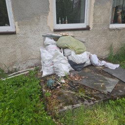 Transporcik - Sterta worków z odpadami budowlanymi przy ścianie budynku z oknami. Widoczne resztki materiałów i gruzu. Trawa i roślinność wokół. Częściowo zdemontowane elementy budynku.