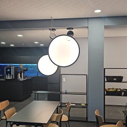 GLE Grzegorz Łachański uslugi elektryczne - Dwie duże, okrągłe lampy wiszące oświetlają strefę jadalnianą z czarnym stołem i drewnianymi krzesłami. W tle nowoczesna kuchnia z ekspresem, a nad całością perforowany sufit.
