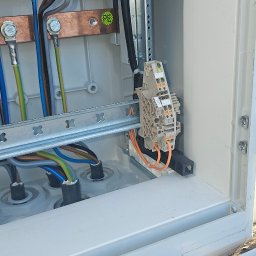 GLE Grzegorz Łachański uslugi elektryczne - Wnętrze otwartej szafki elektrycznej z miedzianymi szynami zbiorczymi, do których podłączono kolorowe przewody. Widoczne terminale na szynie DIN oraz dławiki kablowe. Naklejka 'PE' na szynie.