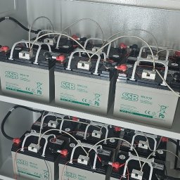 GLE Grzegorz Łachański uslugi elektryczne - Szereg akumulatorów SSB SBL33-12i 12V 33Ah, połączonych białymi przewodami i zabezpieczeniami, zamontowanych na półkach w szarej szafie z wentylacją, tworzących system zasilania awaryjnego.