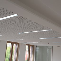 AJ trockenbau - Biały, perforowany sufit podwieszany z wbudowanym oświetleniem liniowym w jasnym pomieszczeniu z oknami. Widoczne elementy instalacji elektrycznej.